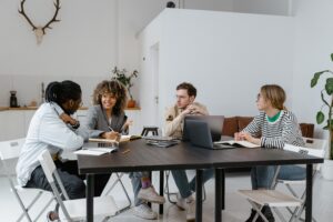 Four people sitting around a table in discussion, with laptops and notebooks open, in a modern office setting.