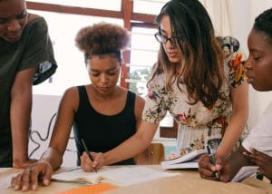 Four young adults working together at a table, writing in notebooks, with one person pointing and guiding the others.