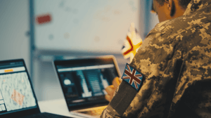 A British soldier in camouflage uniform working on two laptops, displaying maps and data analysis, with a Union Jack patch on their arm, representing involvement in cyber or intelligence operations.
