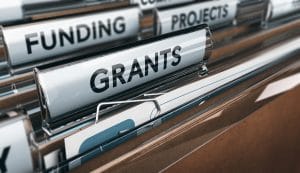 Close-up view of a filing cabinet with folders labelled "GRANTS," "FUNDING," and "PROJECTS." The focus is on the folder labelled "GRANTS.