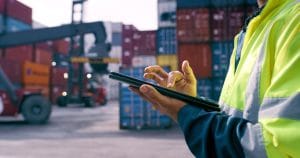 A person wearing a high-visibility yellow jacket is using a tablet device in an industrial shipping yard, with stacked shipping containers and a crane in the background.
