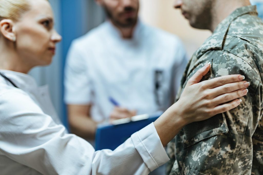 A female healthcare professional places a reassuring hand on a soldier's shoulder during a medical consultation, with another doctor writing notes in the background.
