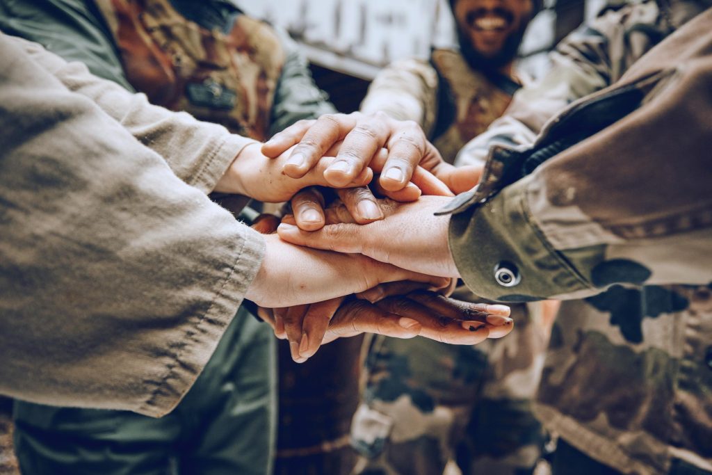 Close-up of diverse soldiers stacking their hands together in a symbol of teamwork and solidarity, smiling and wearing camouflage uniforms.